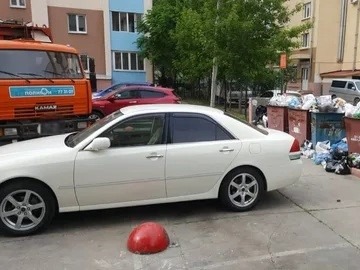 Белый автомобиль припаркован у переполненных мусорных контейнеров во дворе жилого дома.